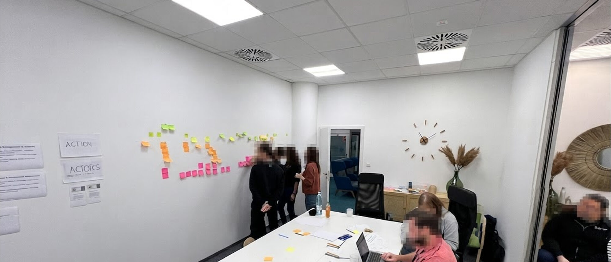 A workshop room with a wall of coloured sticky notes and printed row labels reading ACTION and ACTORS, participants standing at the wall