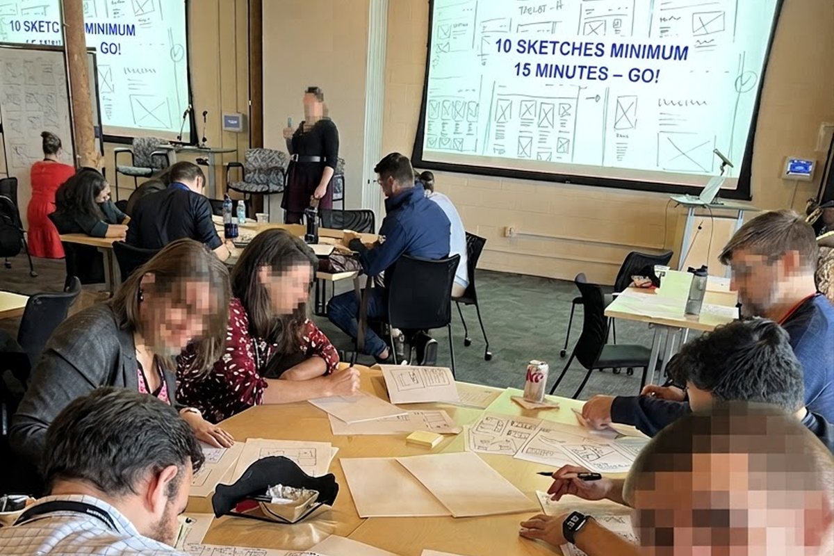 A workshop room mid exercise, the screen reading 10 sketches minimum, 15 minutes, go, participants sketching, faces blurred