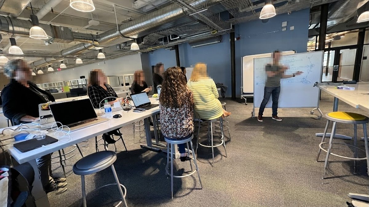 A working session in an open plan office, participants at a long table facing a whiteboard, faces blurred